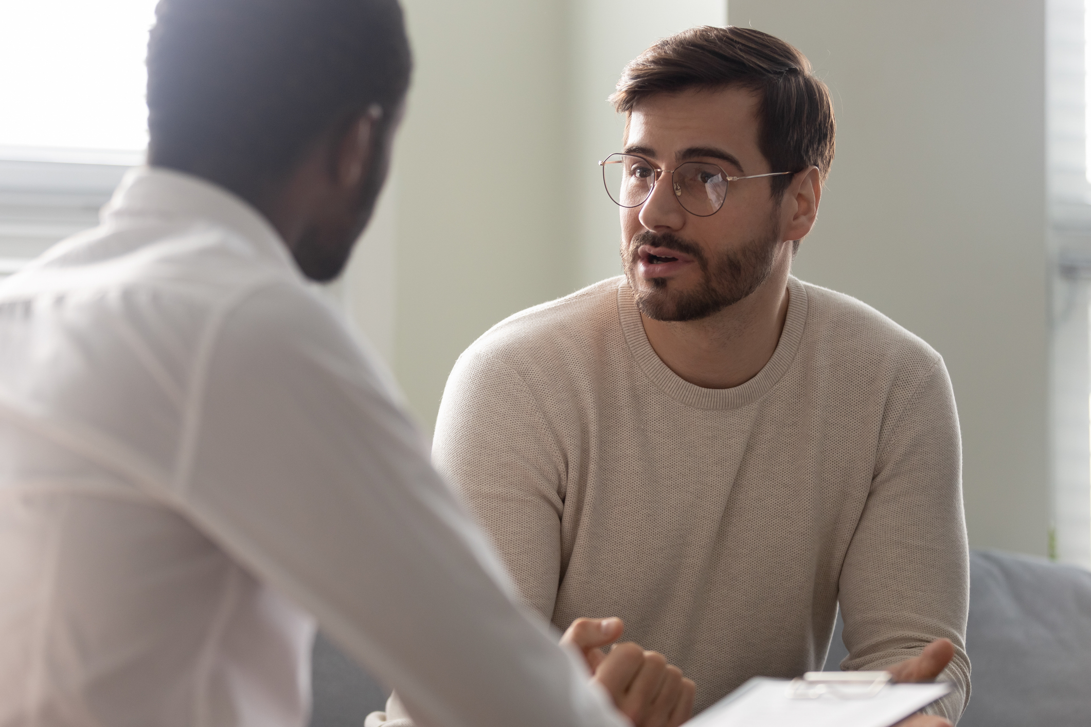 Man consulting with practitioner, African psychologist holding clipboard with card sitting in front of patient listens his mental health complaints. Job interview process applicant and HR manager concept