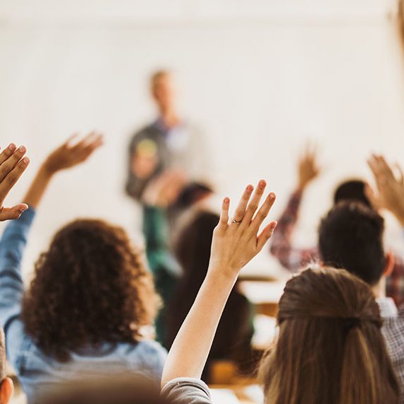 Rear view of large group of students raising their hands to answer the question on a class.